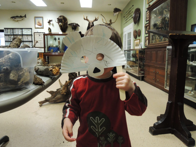 A young child standing in a natural history museum holding a self-made paper mask of a bear over their face.